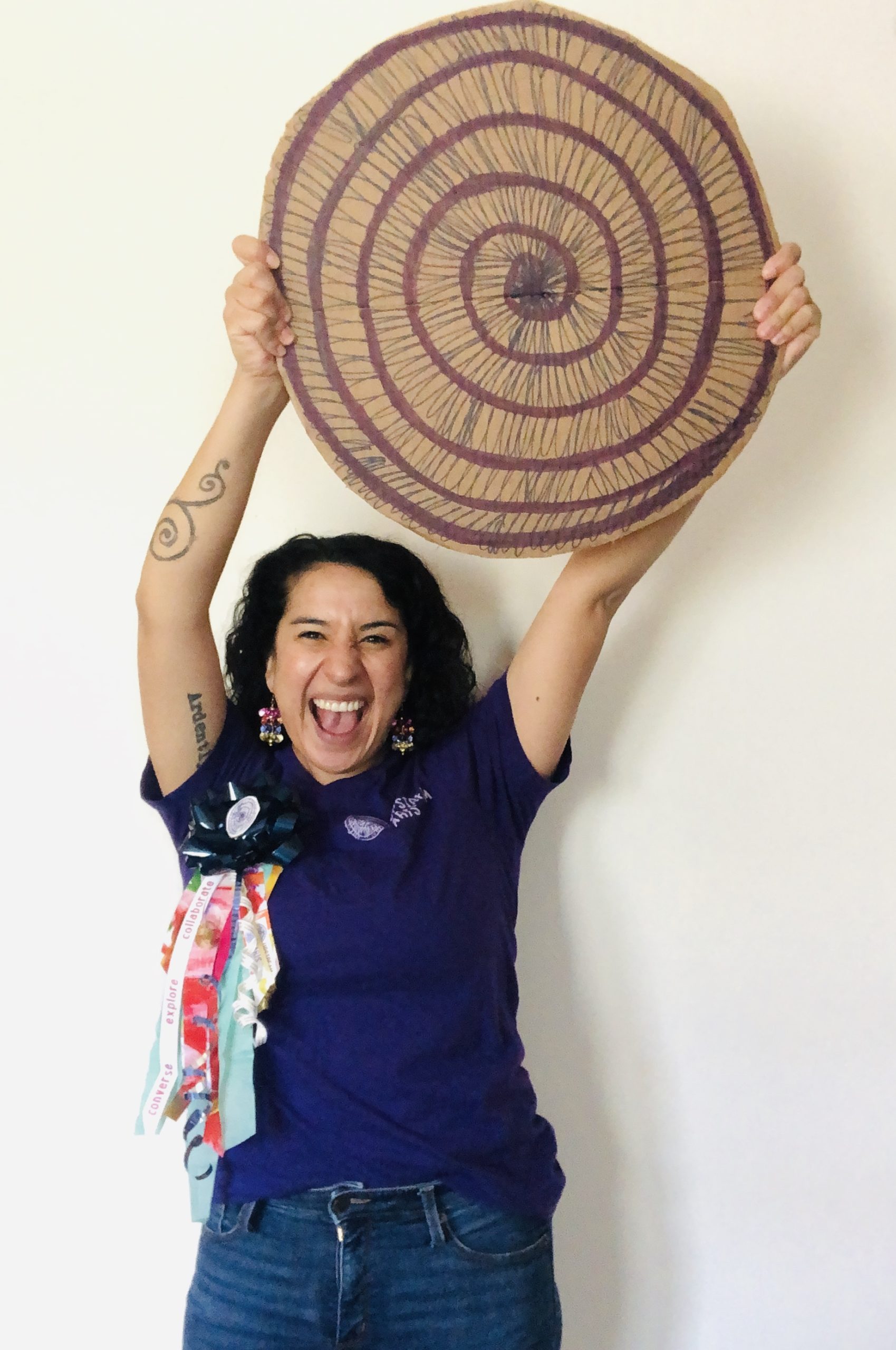 Cristina Castro, Upstream Arts Teaching Artist and At Home Homecoming co-host, wearing a UA t-shirt holds up a homemade UA spiral logo made out of cardboard. On their shirt is a homemade "homecoming mum" from UA annual reports. They are cheering enthusiastically!