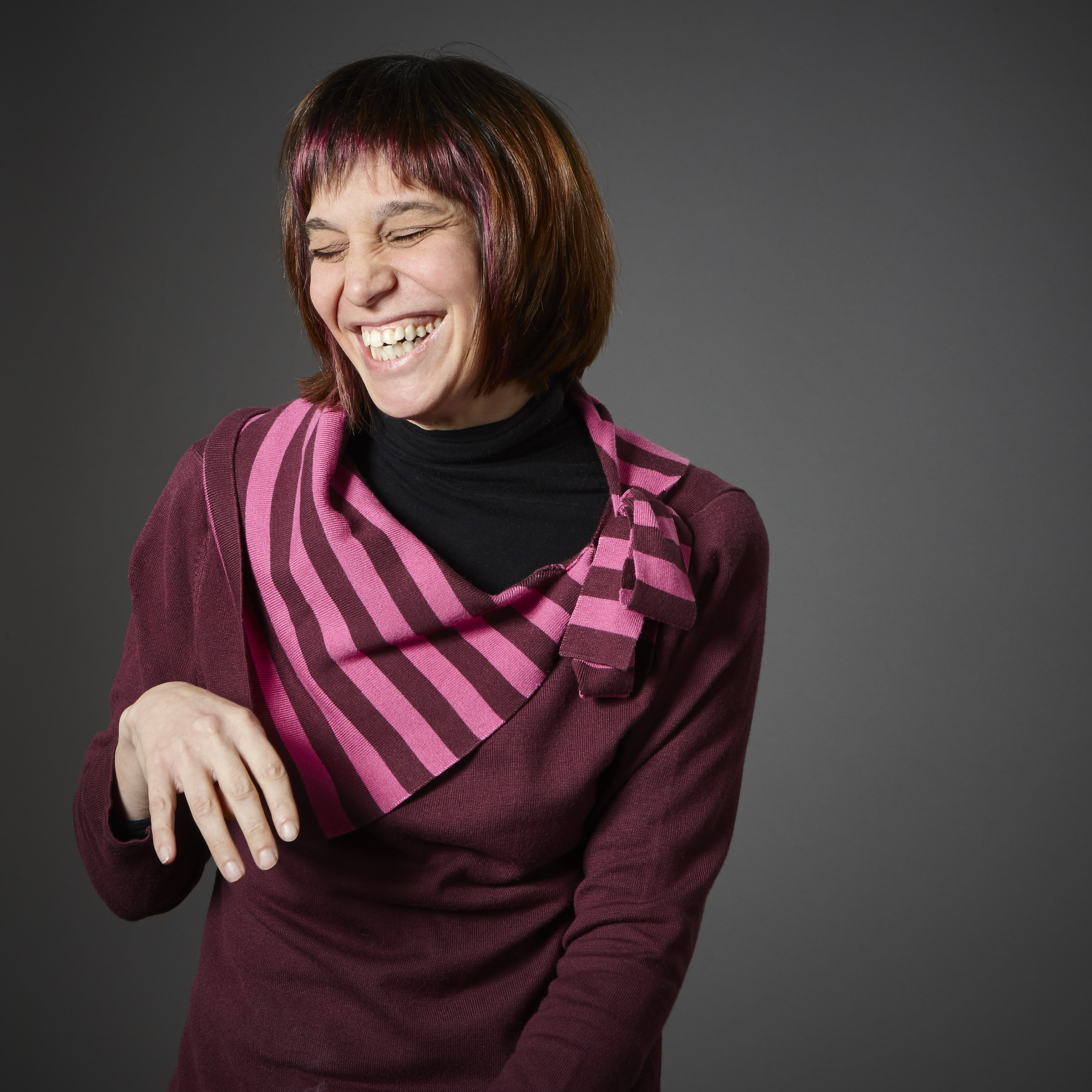 Maria, one of our adult participants, sits for a studio portrait directed by a peer. Her auburn and purple hair, styled in a chin-length bob, beautifully matches her purple striped sweater. She is eyes-closed in a natural smile, looking off slightly to the side.