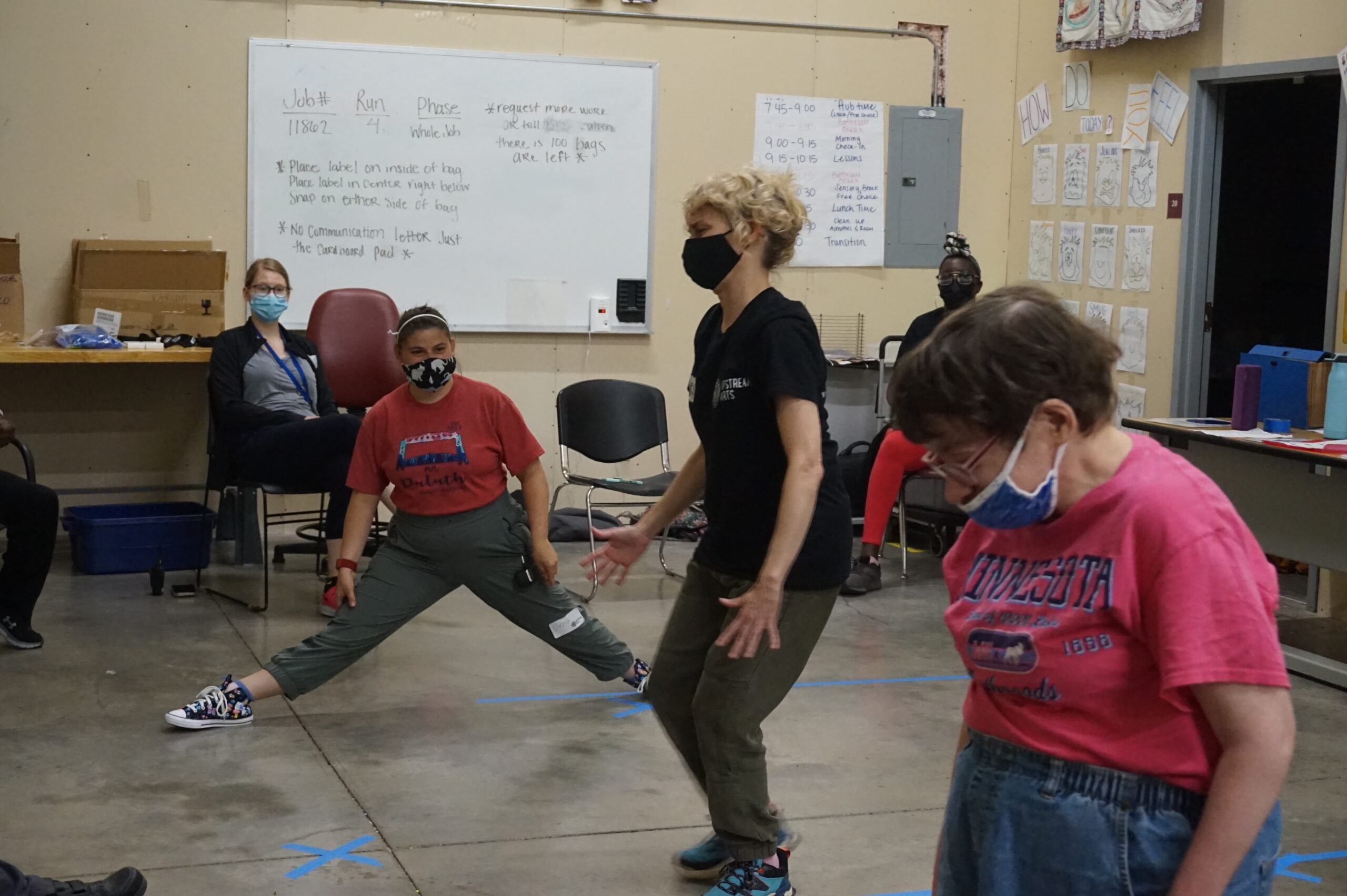 In a classroom, Teaching Artist Tracey, a white woman with blond wavy hair, freezes in place along with two adult participants, one doing the splits and the other looking downwards. Everyone is masked.