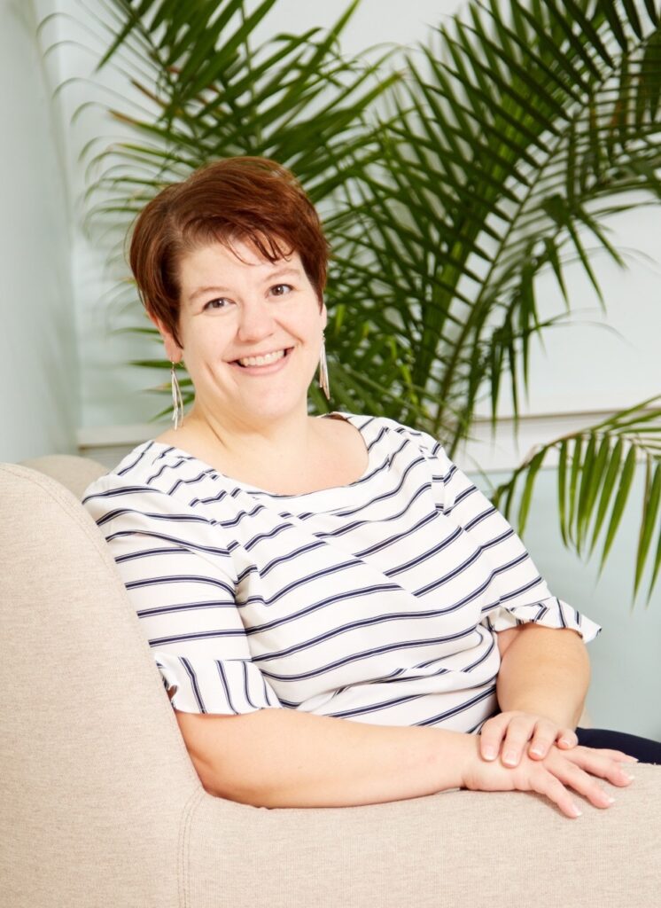Jess Finney is seated in a chair, wearing a white and grey striped shirt, and a warm smile