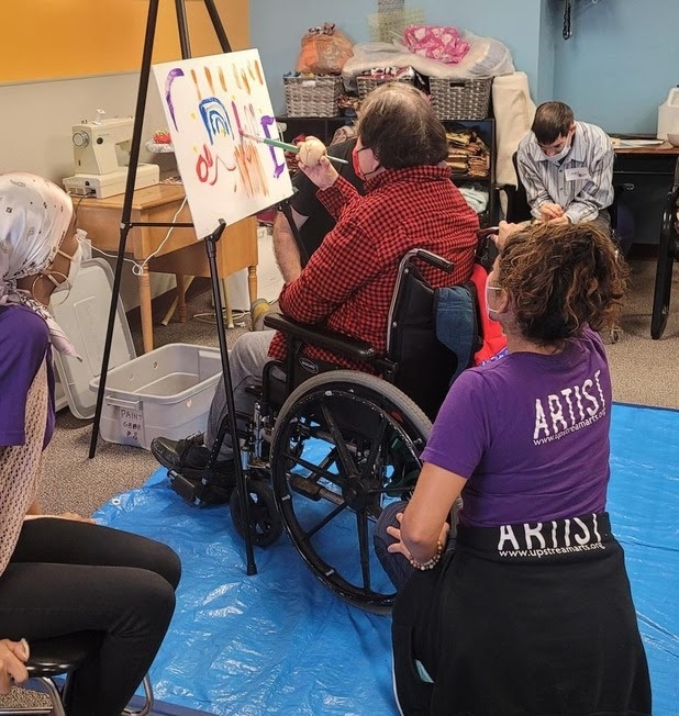 A Participants paints from his wheelchair while a Teaching Artist kneels behind him, watching.